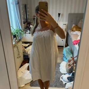 Vit somrig kortklänning med smock - Supersöt vit kortklänning med smala axelband och smock upptill. Klänningen är luftig och har en avslappnad passform, perfekt för varma dagar. Den är ärmlös och har en lätt rynkad volangkant vid bysten för en extra gullig touch.