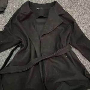 Svart kappa med bälte från Missay - Svart kappa från Missay med klassisk krage och bälte i midjan. Jackan har en stilren och enkel design, perfekt för dig som gillar minimalistiskt mode. Långärmad och i mjukt material som känns skönt mot huden. Snygg att slänga på över vilken outfit som helst.