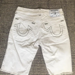 Vita jeansshorts True Religion 32 - Snygga vita jeansshorts från True Religion, modell Rocco Relaxed Skinny. Klassiska bakfickor med kontrastsömmar och råa benslut. Silverfärgade knappar och detaljer, samt tydlig logga bak. Perfekta för en clean och avslappnad look.