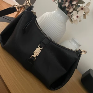 Svart axelväska från Tommy Hilfiger - Snygg svart axelväska från Tommy Hilfiger i skinn med guldfärgade metalldetaljer och dragkedja. Väskan har en stilren design med logga framtill och justerbar axelrem. 