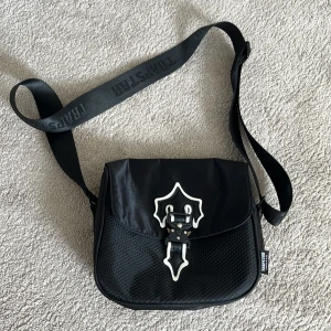 Svart axelväska från Trapstar - Snygg svart axelväska från Trapstar aldrig använd som ny, med bred justerbar rem och coolt spänne framtill. Väskan har mesh-detaljer på framsidan, dragkedja upptill och innerfack med dragkedja inuti