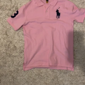 Rosa pikétröja från Ralph Lauren - Säljer en klassisk rosa pikétröja från Ralph Lauren med marinblå logga broderad på bröstet och siffran 3 på ärmen. Tröjan har korta ärmar, krage och knappar framtill. Pris är inte hugget i sten. Passar dig från 170 cm.👌