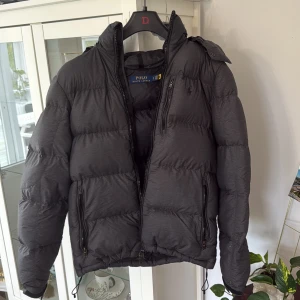 Svart dunjacka från Polo Ralph Lauren - Säljer en svart/grå dunjacka från Polo Ralph Lauren med hög krage som jag köpt från hede fashion outlet i Kungsbacka. Avtagbar huva och flera dragkedjefickor. Jackan har en clean look med RL-detaljer på dragkedjorna och justerbar nederkant. Perfekt för kalla dagar och riktigt snygg till streetwear. Köp på Polo butik för drygt 3800  Priset är inte hugget i sten. 