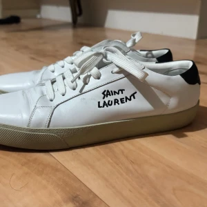 Vita Saint Laurent sneakers 06 i skinn - Snygga vita sneakers från Saint Laurent med svart detalj på hälen och handskriven logga på sidan. Klassisk modell med rund tå, snörning och platt sula. Tillverkade i Italien i äkta skinn, perfekta för en clean och stilren look.