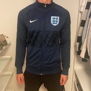Nike England marinblå jacka - Snygg marinblå vindjacka från Nike med Englands landslagsemblem på bröstet. Jackan har hel dragkedja, lång ärm och diskret mönstrad panel över bröst och rygg. Perfekt för fotboll eller chill vardagsstil.