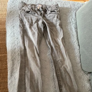 Grå jeans från Gina Tricot - Snygga grå jeansbyxor från Gina Tricot i storlek 152. Byxorna är bootcut, klassisk femficksmodell och coola bakfickor med flätad detalj och knappar. Perfekta för dig som gillar en avslappnad men trendig look.