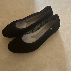 Svarta ballerinaskor från Rieker - Stilrena svarta ballerinaskor från Rieker med rund tå och diskret logga på sidan. Ovandel i mocka och innersula i ljust skinn med blommigt mönster längs kanten. Platt sula med mönster av tassavtryck undertill. Perfekta för en enkel och klassisk look.