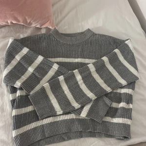 Gråvit randig stickad tröja - Supermysig stickad tröja i grått med vita ränder. Tröjan har en ribbad struktur, lång ärm och en lite högre halsringning. Perfekt för kyliga dagar och passar snyggt till både jeans och kjol.