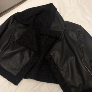 Svart teddyjacka i skinnimitation - Snygg svart teddyjacka med mjukt foder och detaljer i skinnimitation. Jackan har bred krage, dragkedja och en cool, lite boxig passform. Perfekt för dig som vill ha en stilren men edgy look till höst och vinter. Och de är en skinnjacka skulle säga i storlek xs eller s 