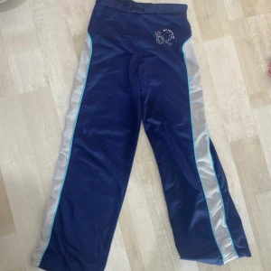 Blå träningsbyxor med silvriga detaljer - Mörkblå träningsbyxor med silvriga meshpaneler längs sidorna och ljusblå piping. I sista bilden ser ni att det är lite defekt.