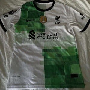 Liverpool Szoboszlai bortatröja Nike - Liverpool FC bortatröja med Szoboszlai 8 på ryggen. Tröjan är vit med gröna grafiska blockdetaljer, svarta tryck och loggor. Klassisk rund hals med svarta muddar. Tillverkad i lätt Dri-FIT-material som andas. Perfekt för dig som vill sticka ut på läktaren eller planen.