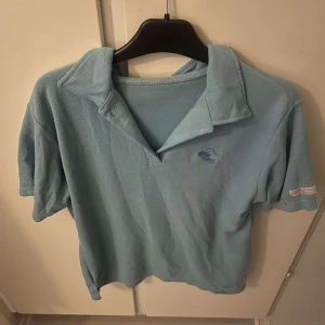 Ljusblå pikétröja från IZOD - Säljer en ljusblå pikétröja från IZOD med klassisk krage och korta ärmar. Tröjan har broderad logga på bröstet och en liten detalj på ärmen. Perfekt för en chill och sportig stil.