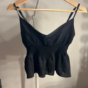 Svart smockad linnetopp med volang - Snygg svart linnetopp med smala axelband och smockad midja som ger en skön passform. Toppen har v-ringning och volang nedtill för en extra touch. Perfekt till jeans eller kjol när du vill ha en enkel men trendig look.