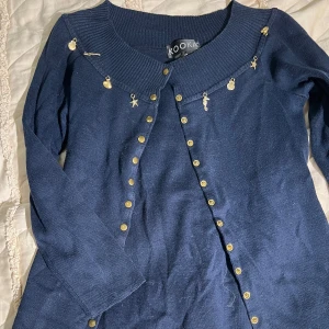 Mörkblå kofta med guldknappar Kookaï - Mörkblå kofta från Kookaï med långa ärmar och dekorativa guldknappar längs hela framsidan. Ribbad krage och unika små guldfärgade berlocker vid halsringningen. Tillverkad i bomull och akryl, perfekt för dig som gillar detaljer.