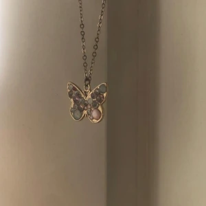 Halsband med fjärilshänge i guld - Supersött halsband med ett fjärilshänge i guldton. Framsidan av fjärilen är dekorerad med små pärlliknande stenar i pastellfärger som rosa, blått och lila. Kedjan är tunn och enkel, perfekt för att ge en gullig touch till din outfit.