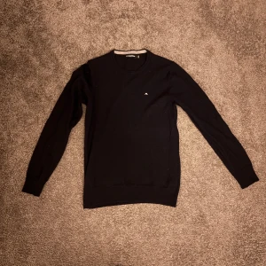 Svart stickad tröja från J.Lindeberg 100% merino - Klassisk svart stickad tröja från J.Lindeberg med rund hals och diskret logga på bröstet. Tröjan har lång ärm och ribbade muddar för en clean look. Perfekt för dig som gillar stilrena och enkla plagg.