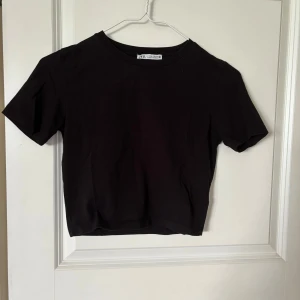 Svart croppad topp från Zara - Enkel och stilren svart croppad topp från Zara med rund halsringning och korta ärmar. Tillverkad i mjuk bomull som känns skön mot huden. Perfekt att matcha med jeans eller kjol för en clean look.