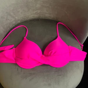 Chockrosa bikiniöverdel med twist - Snygg bikiniöverdel i chockrosa med vadderade kupor och twistad detalj framtill. Smala axelband och enkel knäppning bak. Tillverkad i stretchigt material som sitter skönt och formar sig efter kroppen. Perfekt för stranden eller poolen!