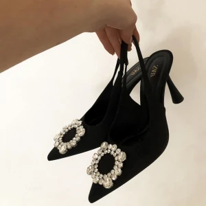 Svarta pumps med strass från Zara - Svarta pumps från Zara med spetsig tå och hög klack. Skorna har en snygg rem runt hälen och är dekorerade med stora, glittrande strassdetaljer framtill. Tillverkade i ett matt tyg som ger en lyxig känsla och passar perfekt när du vill sticka ut. Använd endast en gång så de är i mycket bra skick.