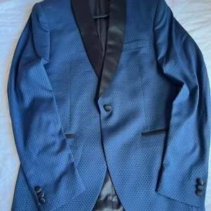 Blå mönstrad kavaj Isaac Dewhirst - Snygg blå kavaj från Isaac Dewhirst med små diskreta prickar och svarta satindetaljer på slag och fickor. Kavajen har två knappar framtill och knappar vid ärmsluten. Perfekt för dig som vill sticka ut med stilren look.