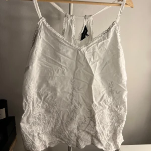 Vit broderad linnetopp från New Look - Superfin vit linnetopp från New Look med tunna axelband och v-ringning. Toppen har broderade blommor nertill och spetsdetaljer längs kanterna. Perfekt för varma dagar och ger en romantisk vibe. Bara att skriva vid frågor!🩷