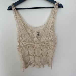 Beige spetslinne från Vero Moda  - Supersött beige linne från Vero Moda Jeans i storlek XS. Toppen är virkad med blommiga spetsdetaljer och har en croppad passform. Perfekt för varma dagar eller lager på lager. Snyggt mönster och fransig nederkant ger en boho-vibe. Använd fåtal gånger