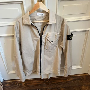 Beige overshirt från Stone Island - Snygg beige overshirt från Stone Island med klassisk logga på ärmen. Jackan har dragkedja framtill, bröstficka med knapp och en clean, minimalistisk look. 