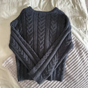 Mörkblå stickad tröja med flätmönster - Mysig mörkblå stickad tröja med klassiskt flätmönster över hela plagget. Tröjan har rund halsringning och långa ärmar, perfekt för kyliga dagar. Den är mjuk och passar dig som gillar en enkel men ändå stilren look.