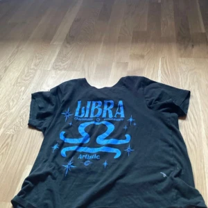 Svart Libra t-shirt från Gina Tricot - Cool svart t-shirt från Gina Tricot med blått Libra-tryck och stjärndetaljer på ryggen. Oversized fit med korta ärmar och rund hals. Perfekt för dig som gillar astrologi och vill sticka ut med en unik printad tee.