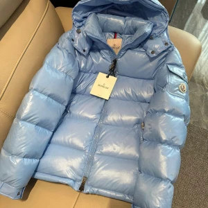 Moncler Maya - Snygg ljusblå dunjacka från Moncler med glansig finish och huva. Jackan har dragkedja framtill, ficka med lock på ärmen och klassisk Moncler-logga. Perfekt för kalla dagar när du vill hålla stilen och värmen. Kontakta via Snapchat: masionla_drippo för eventuell rabatt på denna produkt!