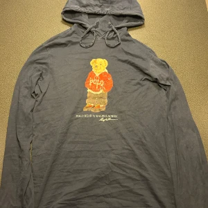 Mörkblå hoodie med Polo Bear från Ralph Lauren - Säljer en mörkblå hoodie från Ralph Lauren med den ikoniska Polo Bear-trycket på bröstet. Tröjan har långa ärmar, och är tillverkad i mjuk bomull. Perfekt för en avslappnad och cool stil. Vid frågor eller funderingar är de bara att höra av sig!