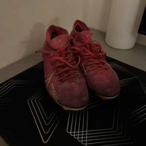 Rosa fotbollsskor från Nike - Säljer ett par rosa Nike fotbollsskor med gula detaljer och snörning. Skorna har en smal passform och är designade för spel på konstgräs eller gräs. Yttermaterialet är syntet och sulan är gul med dobbar för bra grepp.
