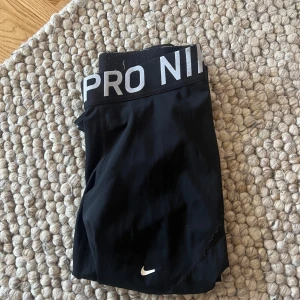 Svarta träningstights från Nike Pro - Svarta Nike Pro tights med bred resår i midjan där det står 'NIKE PRO'. Tillverkade i Dri-FIT-material som andas och transporterar bort fukt. Perfekta för träning och sportaktiviteter.