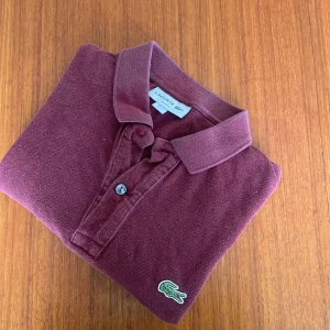 Vinröd Lacoste piké - Stilren vinröd Piké från Lacoste. Väldigt bekväm. Är perfekt för en varm sommardag.  Skicket är 9/10 pågrund av att knappen högst upp vid kragen saknas.
