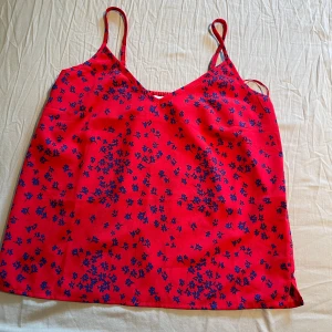 Röd blommig linnetopp från SAMSA Collection - Säljer en röd linnetopp från SAMSA Collection med tunna axelband och blått blommönster. Toppen har en lös passform och är perfekt för varma dagar. Superfin att styla med jeans eller kjol!