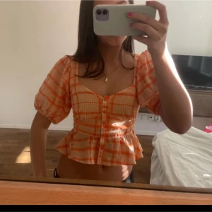 Orange och aprikosfärgad blus med puffärmar - Superfin blus i orange och aprikos med rutigt mönster. Toppen har korta puffärmar, knappar framtill och en volang nertill. Snygg fyrkantig ringning och croppad modell som passar perfekt till sommaren. Står ingen storlek men passar S och XS 