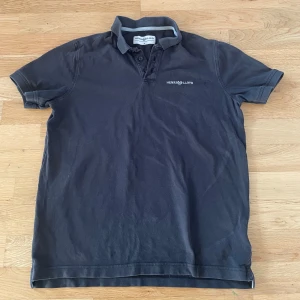 Pikétröja från Henri Lloyd - Säljer en svart pikétröja från Henri Lloyd med klassisk krage och knappar framtill. Tröjan har korta ärmar och diskret logga broderad på bröstet. Perfekt för en stilren och avslappnad look.