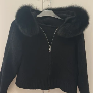Svart stickad jacka med fuskpäls - Snygg svart stickad Alessa jacka med dragkedja och stor huva kantad med mjuk fuskpäls. Jackan har ribbade muddar vid ärmslut och nederkant. Perfekt för en trendig look. Nypris 1800kr
