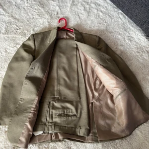 Beige kostym med kavaj och byxor - Säljer en klassisk beige kostym med matchande kavaj och byxor. Kavajen har tre knappar framtill, spetsiga slag och flera fickor. Kostymen är helfodrad och har en stilren, tidlös design som passar till många tillfällen.