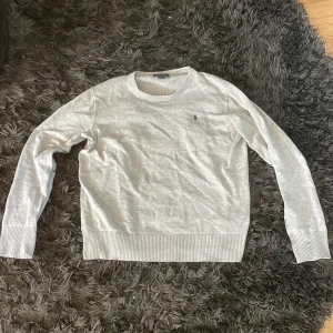 Ljusgrå tröja från Polo Ralph Lauren - Säljer en stilren ljusgrå tröja från Polo Ralph Lauren med rund halsringning och diskret broderad logga på bröstet. Tröjan har långa ärmar och ribbade muddar vid ärmslut och nederkant. Perfekt för en klassisk och avslappnad look.
