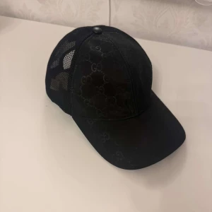 Svart keps från Gucci med mesh - Säljer en svart keps från Gucci med diskret GG-mönster på framsidan och mesh på sidorna och baksidan. Justerbar storlek baktill med plastspänne. Perfekt för dig som gillar stilrena accessoarer med lyxig känsla. Gucci påse ingår
