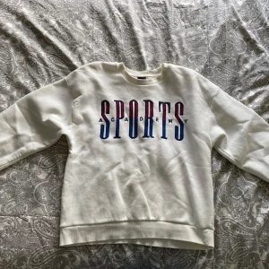 Vit sweatshirt med tryck från Gina Tricot - Säljer en vit sweatshirt från Gina Tricot med stort blått och rött tryck där det står 'SPORTS ACADEMY' på bröstet. Tröjan har rund halsringning och långa ärmar. Perfekt för en avslappnad stil.