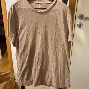 Beige t-shirt i bomull - Säljer en enkel beige t-shirt i mjuk bomull. T-shirten har rund halsringning och korta ärmar. Perfekt för en avslappnad stil och lätt att matcha med andra plagg. Ett hål på, litet.