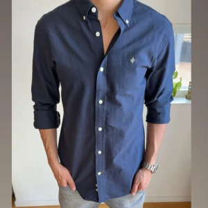 Morris skjorta - Säljer en stilren mörkblå skjorta (knappast använd) från Morris med vit broderad logga på bröstet. Skjortan har klassisk krage, vita knappar och lång ärm. Perfekt för dig som gillar en clean och tidlös look. Den är i M 39/40.