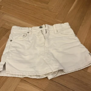 Vita jeansshorts från Zara - Säljer ett par vita jeansshorts från Zara med klassisk femficksdesign och knappgylf. Shortsen har bälteshällor och raka ben med små slitsar i sidorna. Perfekta för varma dagar och enkel att matcha med olika toppar. 