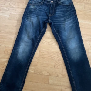 Blå jeans från SMOG - Säljer ett par klassiska blå jeans från SMOG med snygga slitningar och raka ben. Jeansen har fem fickor, orangea sömmar och normal passform. Perfekta till vardags för en avslappnad stil.