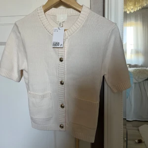 Vit stickad kofta från H&M - Säljer en söt vit stickad kofta från H&M med korta ärmar och ribbade kanter. Koftan har knappar framtill och två fickor. 