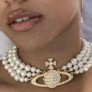 Pärlhalsband med ikonisk hänge från Vivienne Westwood - Lyxigt halsband med tre rader vita pärlor och ett stort, glittrigt guldfärgat hänge i form av Vivienne Westwoods klassiska orb-symbol, dekorerat med gnistrande stenar. Perfekt statement piece för att lyfta din stil.