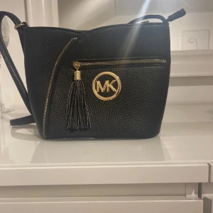 Svart axelväska från Michael Kors - Säljer en svart axelväska från Michael Kors med guldfärgade detaljer, dekorativ tofs och MK-logga framtill. Väskan har en synlig dragkedja och är tillverkad i strukturerat skinn. Perfekt för dig som vill ha en stilren och trendig accessoar. Fick den i present så antar att den är äkta men är inte säker 