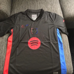 FC Barcelona svart bortatröja 24/25 - Säljer en svart FC Barcelona bortatröja från Nike med röda och blå detaljer på sidorna. Tröjan har klubbmärke och Nike-logga på bröstet samt Spotify som sponsor. Tillverkad i Dri-Fit material för bra komfort.